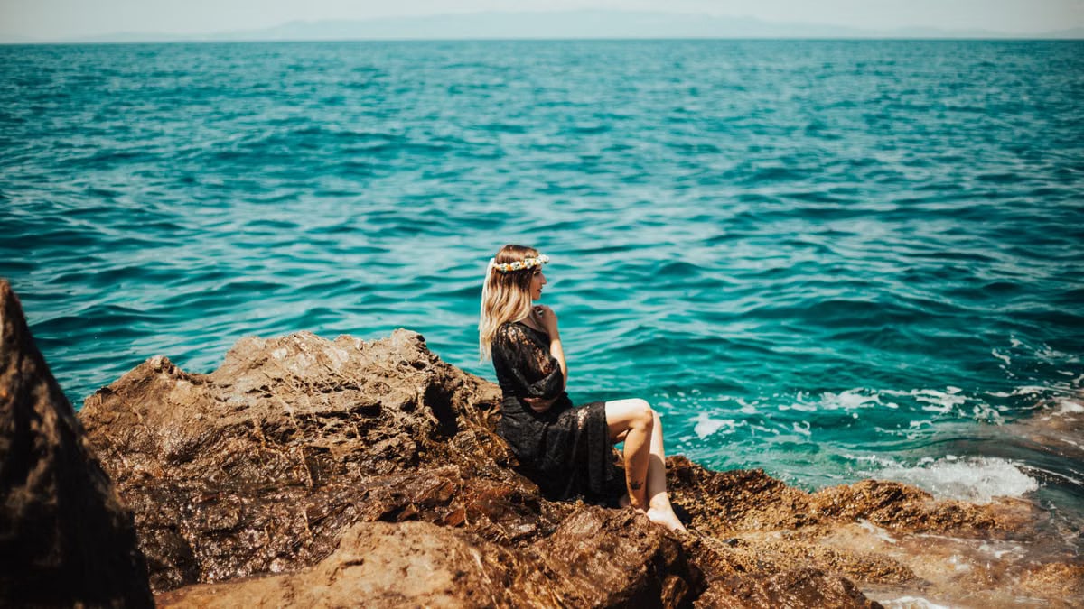 A woman in a black dress and floral wreath sits on rocks by the sea in İzmir, Turkey.